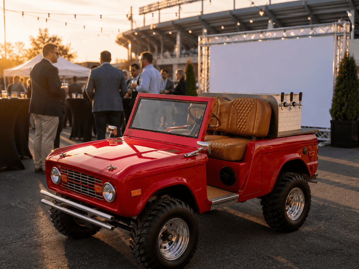 Red vintage Bronco-style bar cart at a stadium-side corporate event at golden hour, suited guests networking at cocktail tables under string lights