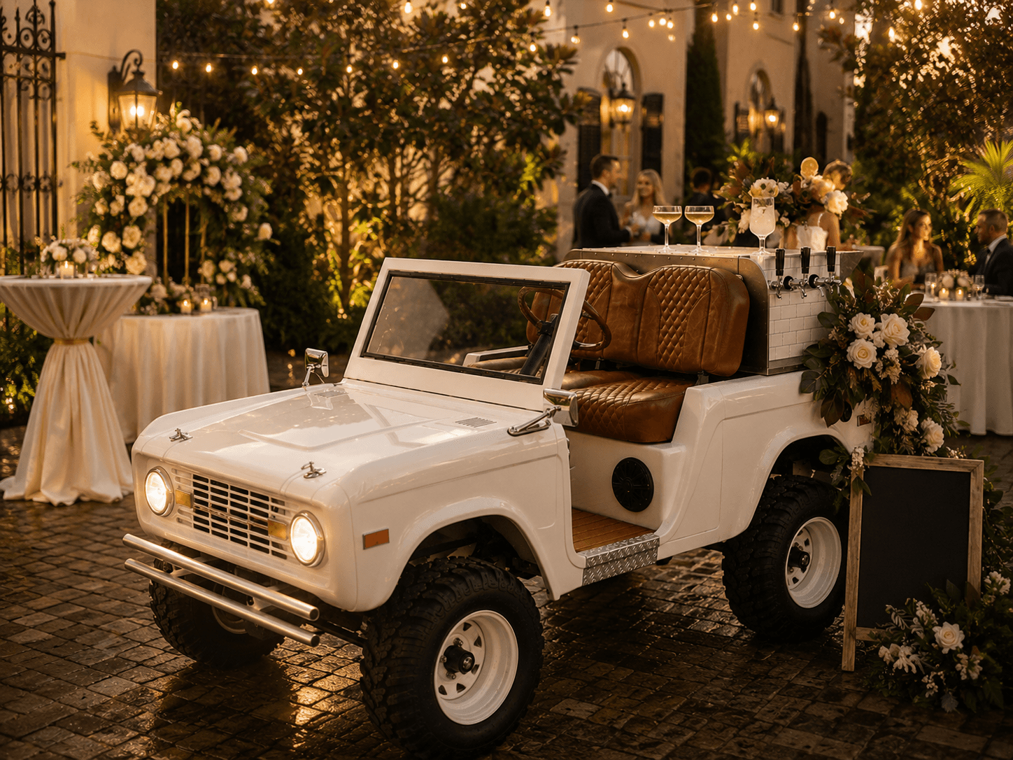 White vintage Bronco-style bar cart styled for a courtyard wedding — florals, gas lanterns, cobblestones, bride and guests in the background at golden hour