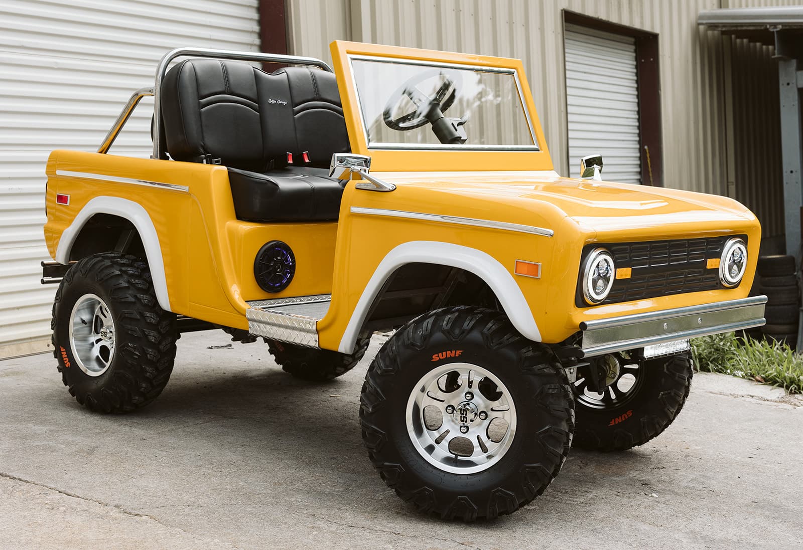Yellow Vintage Bronco cart with black seats and off-road tires, parked outside the shop